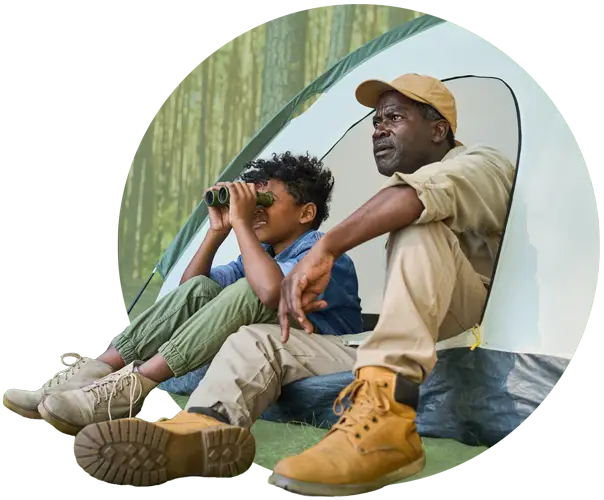 Photo of a child looking through binoculars sitting beside an older man. Both are sitting inside a tent with their legs outside of it.