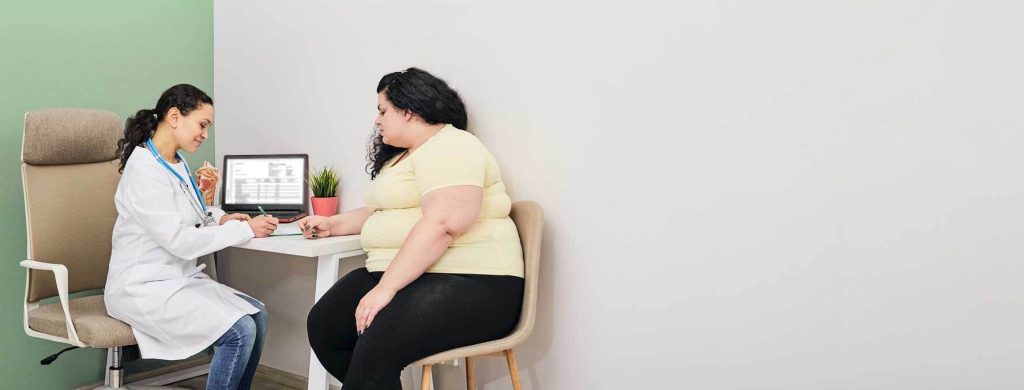Photo of a heavyset woman sitting at a desk and speaking with a healthcare provider taking notes
